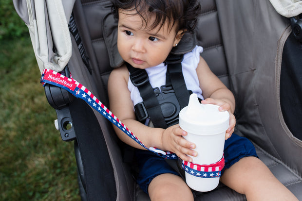 Toddler using BooginHead SippiGrip in American Flag to keep the sippy cup clean and attached to their stroller. Size adjustable strap can attach to bottles, sippy cups or toys. Essential baby and toddler supply! Easy to clean. 