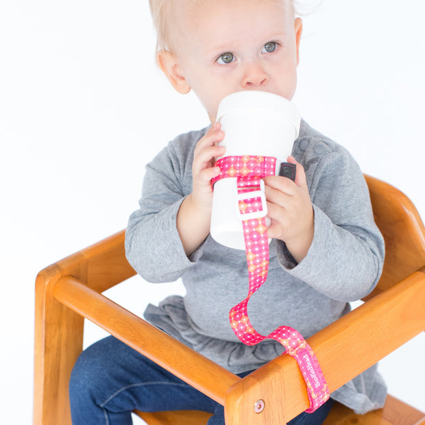 Toddler using BooginHead SippiGrip in Dottie, hot pink and white design, to keep the sippy cup clean and attached to their highchair. Adjustable strap can attach to bottles, sippy cups or toys. Essential baby and toddler supply! Easy to clean. 