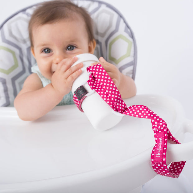 Toddler using BooginHead SippiGrip in Pink Polka Dots to keep the sippy cup clean and attached to their highchair. Size adjustable strap can attach to bottles, sippy cups or toys. Essential baby and toddler supply! Easy to clean. Bright pink and white design